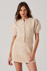 A woman is wearing a light cream-colored, knitted two-piece outfit. The top is a short-sleeved cardigan with a round neckline, buttoned down the front, featuring puffed sleeves and decorative pearl embellishments. The skirt is a matching mini skirt, also detailed with pearl embellishments, creating a coordinated, textured look.